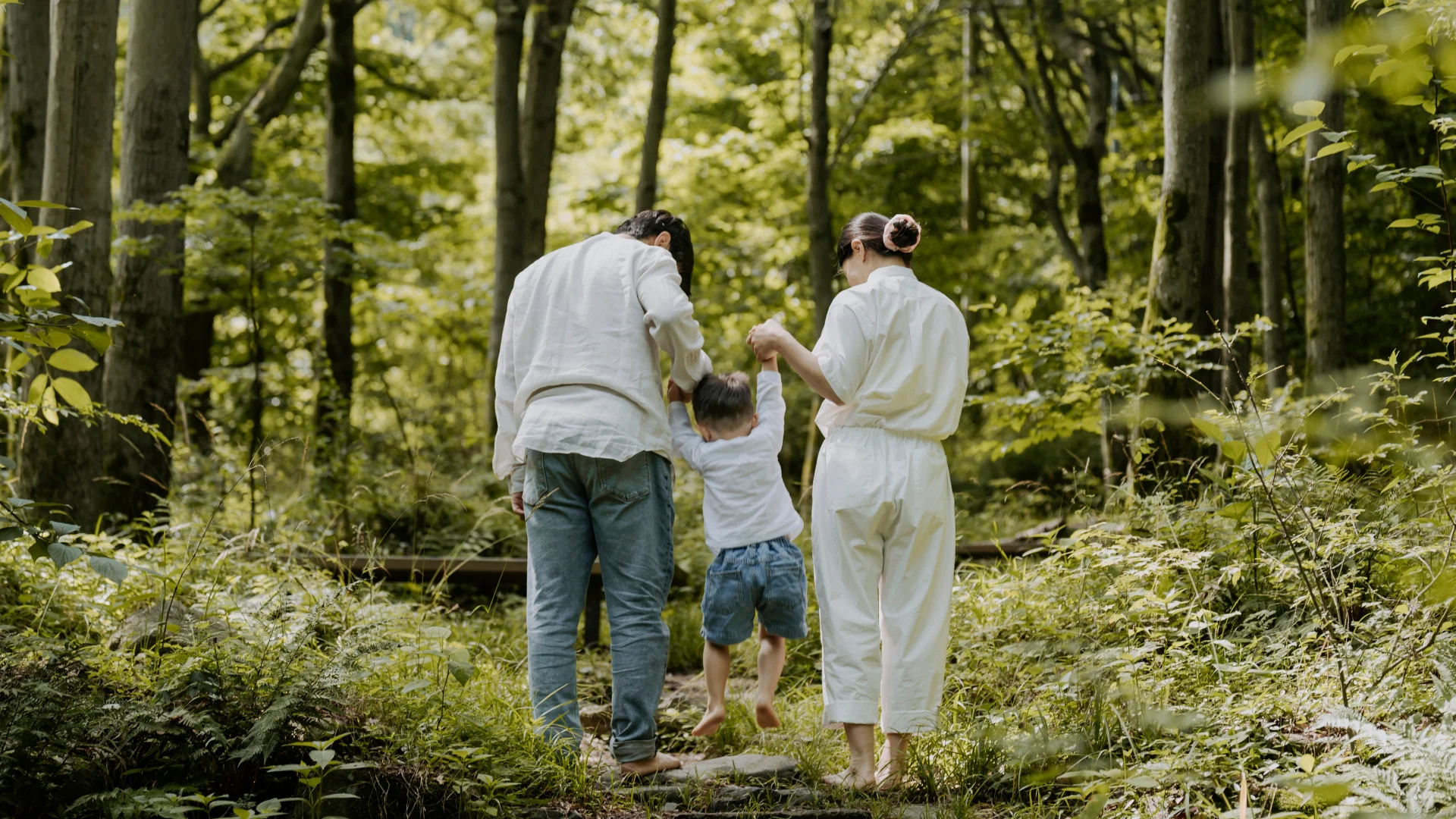This image shows two adults and a child walking together through a sunlit forest, holding the child's hands as they explore the lush green woodland.