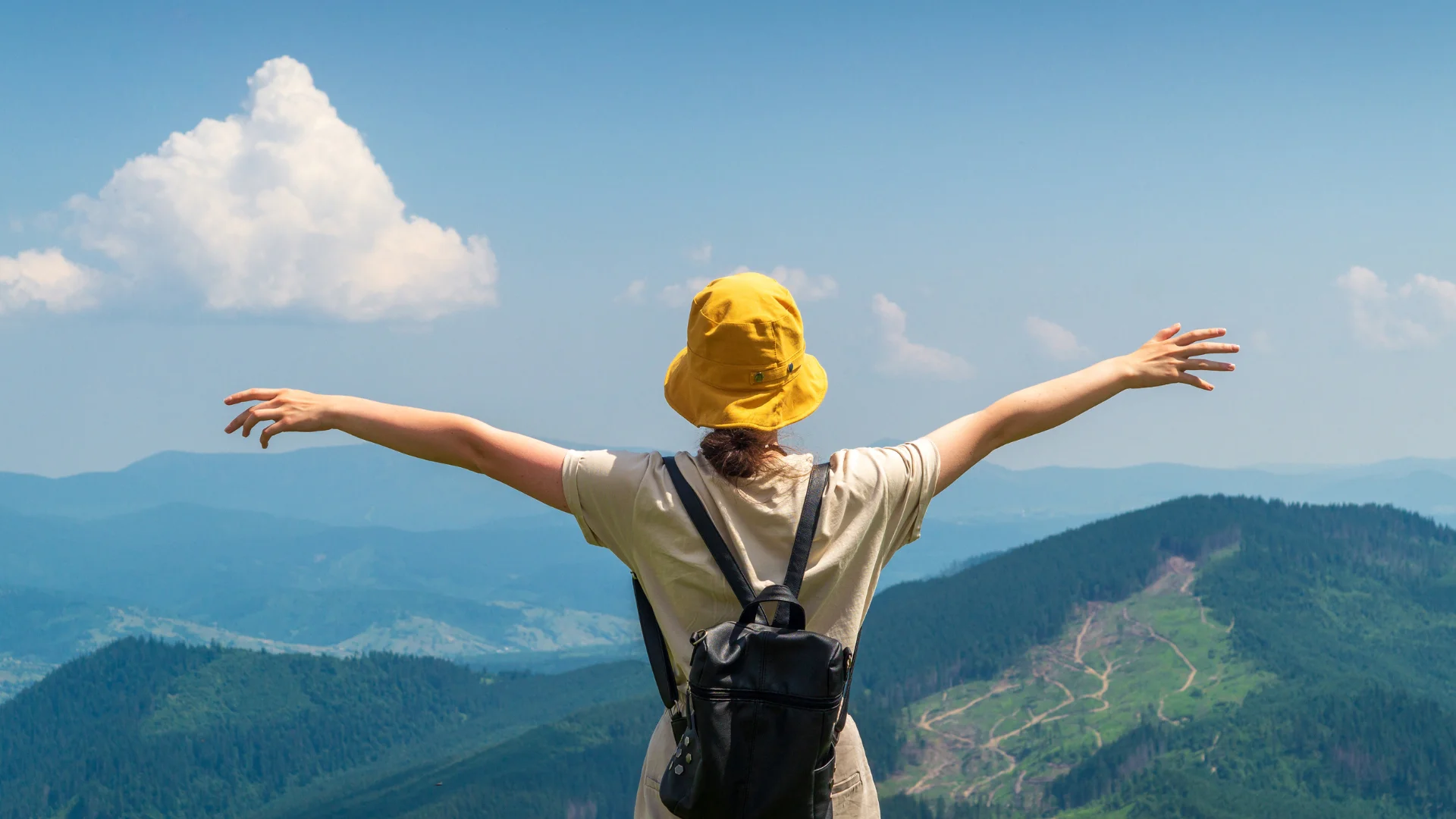 This image shows a person wearing a yellow hat with arms outstretched, viewing a panoramic mountain landscape from a high vantage point under a blue sky.